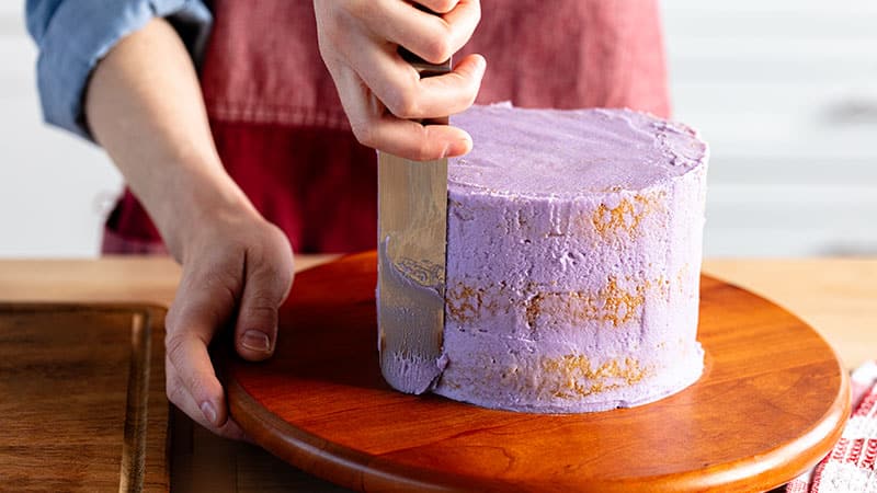  a person smoothing out purple frosting on a 3 layer cake atop a wood cake wheel