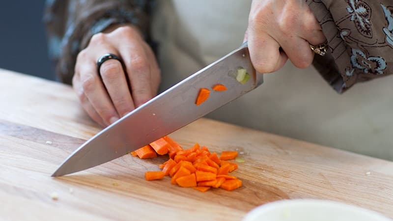 Hands and knife chopping carrots on a butcher block table.