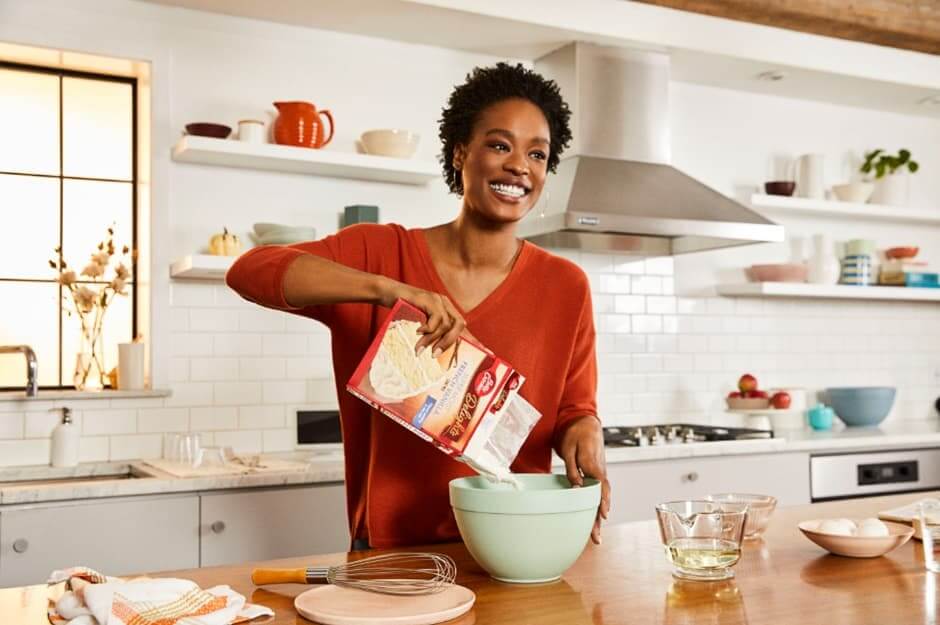 A smiling woman in a v-neck top pours cake mix into a bowl in a bright kitchen setting, with baking tools on the counter.